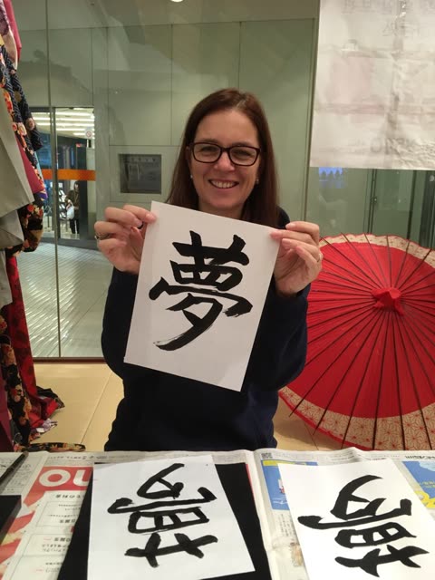 A woman in a blue shirt points to a Japanese character on paper for another person holding a calligraphy brush at a table.