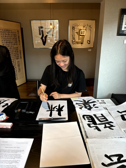 A woman in a blue shirt points to a Japanese character on paper for another person holding a calligraphy brush at a table.