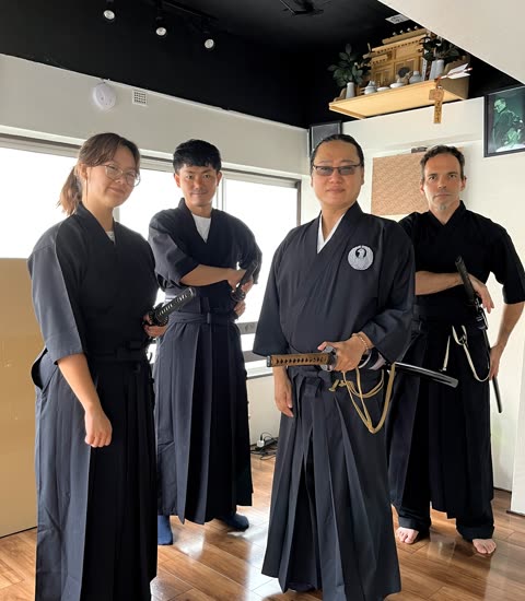 Person in black kimono handling a sword near a sword rack.