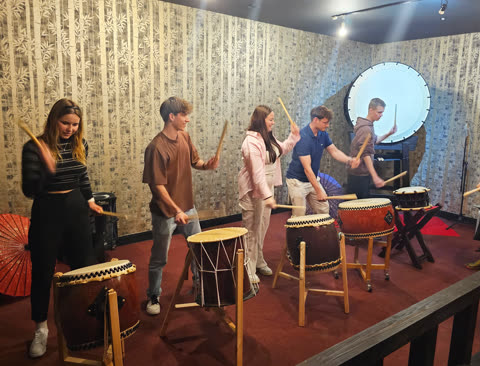 Four people stand holding large drumsticks in front of individual taiko drums in a practice room.