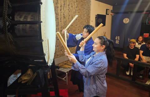 Four people stand holding large drumsticks in front of individual taiko drums in a practice room.