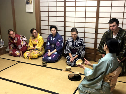 Five people in colorful kimonos kneel on a tatami mat floor, drinking from small bowls in front of a shoji screen.