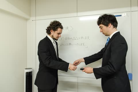 Two men in suits exchange business cards in front of a whiteboard.