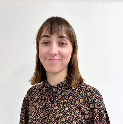 A woman with brown shoulder-length hair and bangs wearing a patterned shirt smiles in front of a white wall.
