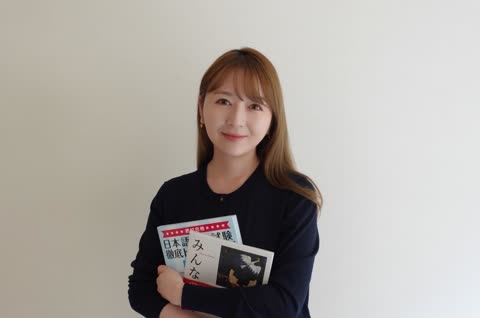 A woman with light brown hair holds two Japanese textbooks while standing against a plain wall.