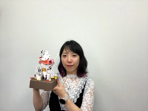 A woman with dark hair holds a decorative object resembling a parfait with paper cutouts against a white wall.