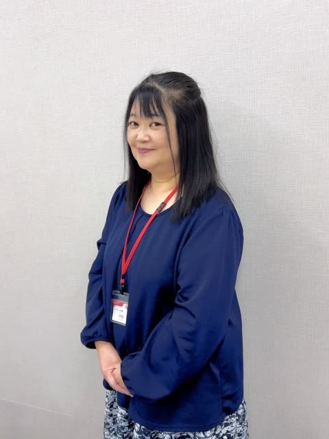 An East Asian woman with shoulder-length black hair stands with her hands clasped in front of a light gray textured wall.