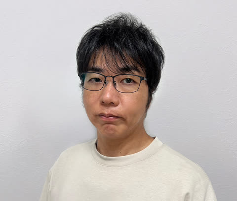 A man with dark hair and glasses wearing a light-colored t-shirt looks at the camera against a plain white wall.