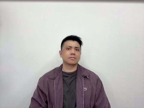 A person in a purple jacket over a dark t-shirt looks at the camera while standing against a white wall.